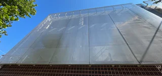 Moderne Gebäude-Fassade mit transparenter Metallgewebe-Verkleidung von Haver & Boecker unter blauem Himmel, teils von Bäumen umrahmt.